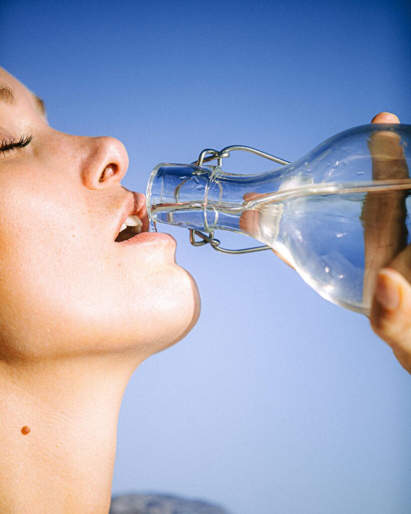 A woman drinking water from a glass bottle against a clear blue sky, emphasizing hydration.